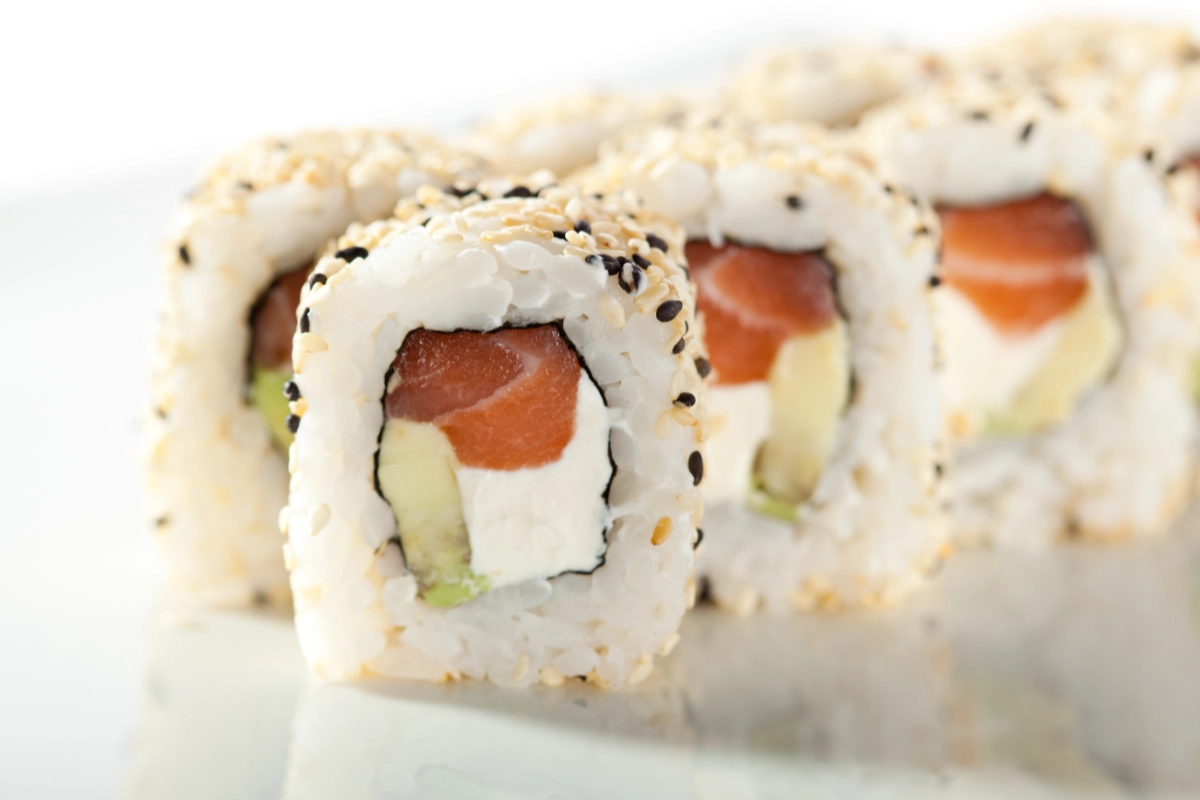 Close-up of an Alaska sushi roll, featuring rice with sesame seeds, avocado, and salmon, on a white background.