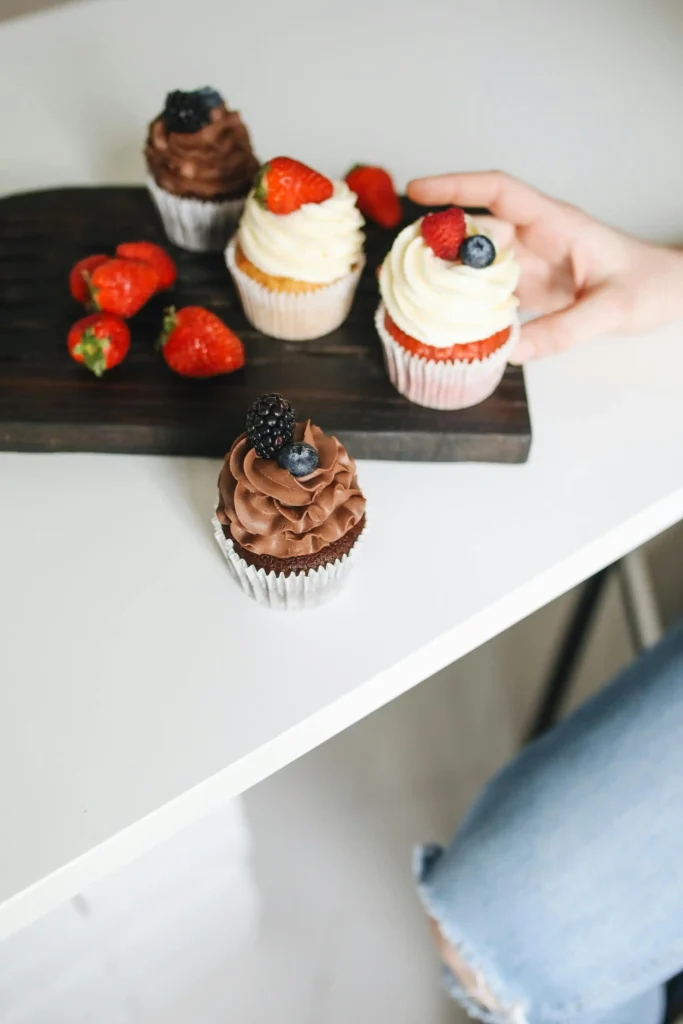 A hand is picking a gluten-free cupcake topped with a blackberry and blueberry from a wooden board holding various cupcakes, some topped with strawberries, against a white countertop background.