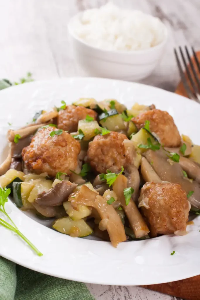 A plate of oyster mushrooms stir-fried with chunks of chicken and garnished with parsley, with a bowl of white rice and a fork in the background.