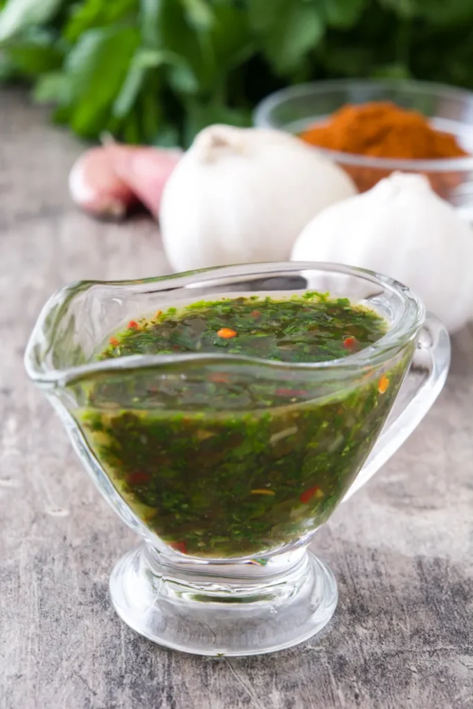 A clear glass gravy boat filled with cilantro chimichurri sauce, with fresh herbs visible, on a wooden surface. In the blurred background, there are garlic bulbs and a small bowl of red spice.