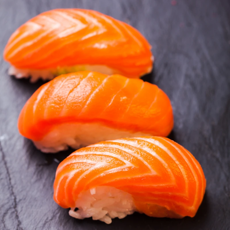 Three pieces of sake sushi, with lustrous salmon atop compact rice, arranged in a line on a dark slate surface.