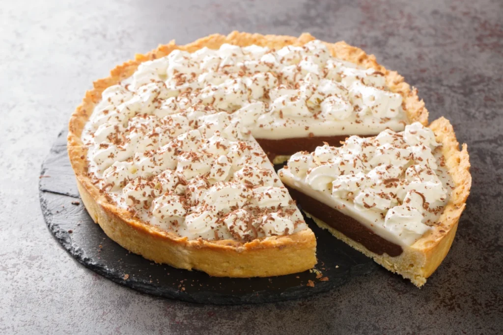 A haupia pie with a golden-brown crust, topped with whipped cream and sprinkled with grated chocolate, is partially sliced on a dark plate with a light dusting of chocolate.