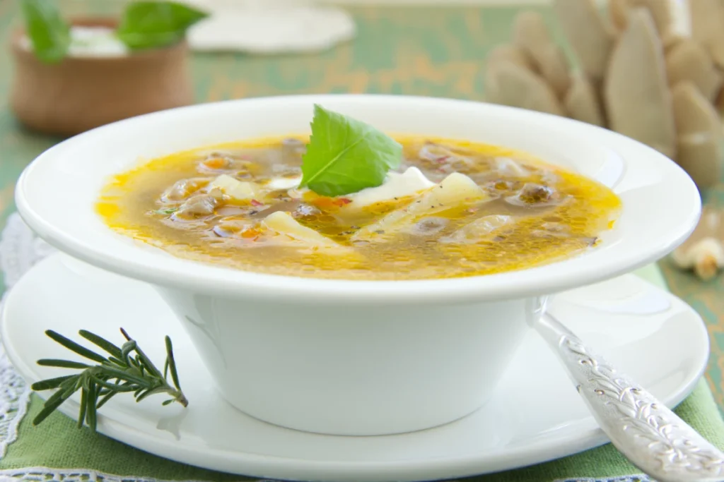 A bright bowl of oyster mushroom soup with a dollop of sour cream and a basil leaf garnish, served on a white plate with a decorative spoon, on a green tablecloth. Sprigs of rosemary and whole mushrooms are in the background.