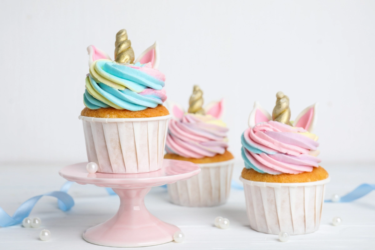 Three cupcakes decorated to resemble unicorns, with swirls of pastel blue and pink frosting and golden horns, displayed on white and pink cake stands.