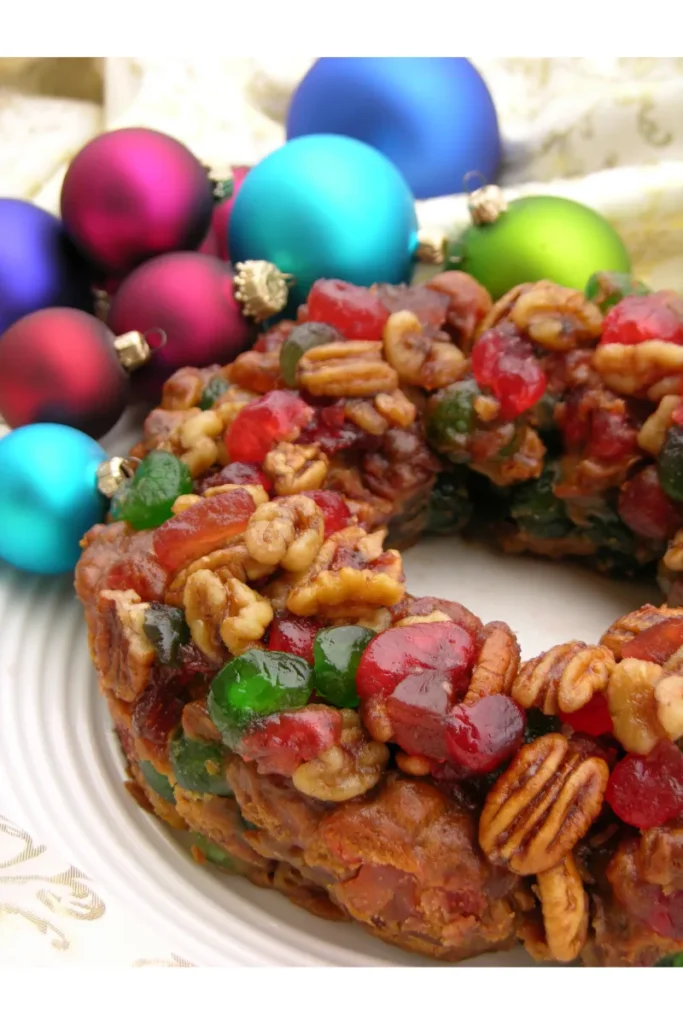 Close-up of a fruitcake with abundant candied fruits and pecans on top, with a backdrop of colorful Christmas ornaments in red, blue, and green, suggesting a festive holiday setting.