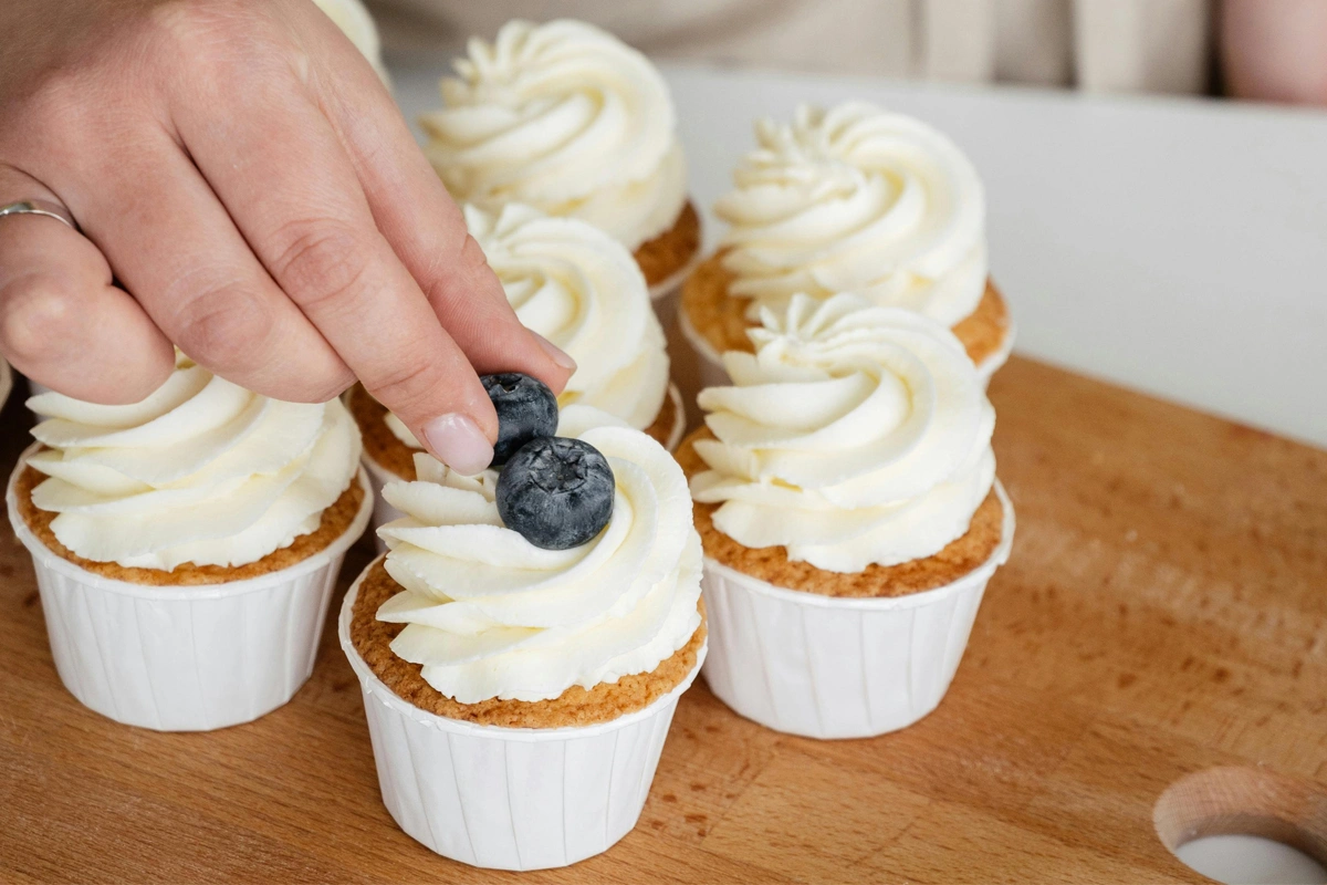 A hand places a blueberry atop a gluten-free cupcake with creamy white frosting, arranged with other similar cupcakes on a wooden surface.