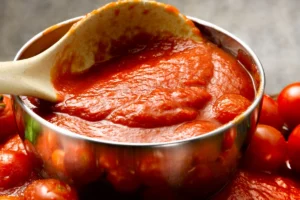 A close-up view of a pot of thick tomato gravy with a wooden spoon lifting some out, surrounded by whole tomatoes, emphasizing the fresh ingredients.