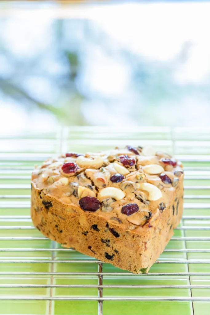 A loaf of fruitcake with nuts and dried fruits on top, cooling on a wire rack with a blurred green background.