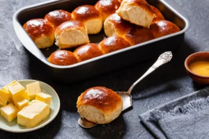 A tray of golden-brown Hawaiian rolls in a baking dish, with a plate of butter cubes and a small bowl of melted butter beside it, ready for serving.