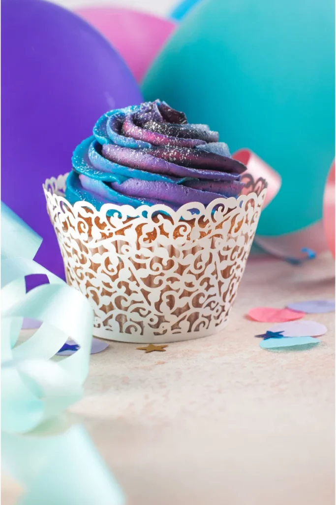 A cupcake with blue and purple galaxy frosting and sparkling edible glitter is nestled in a decorative white wrapper. In the soft-focus background, pastel balloons and ribbons contribute to a festive ambiance, with a few star-shaped confetti pieces in the foreground.