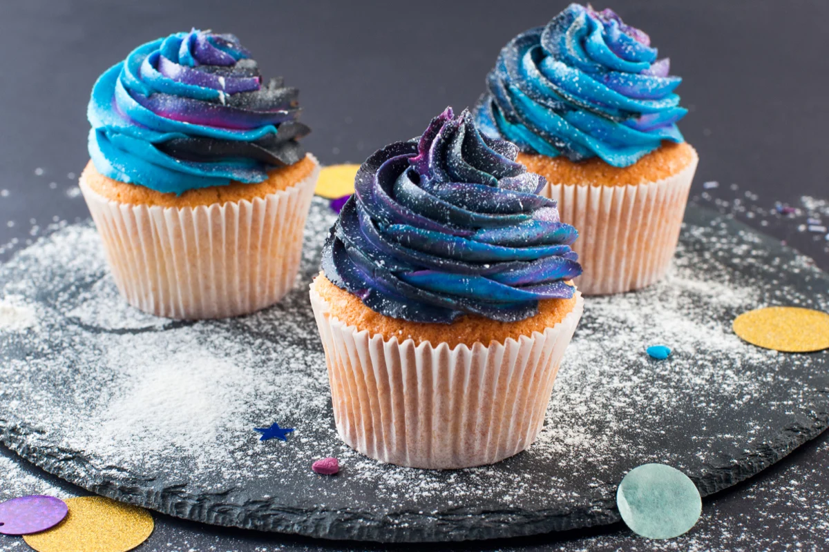 Three cupcakes with galaxy-themed frosting in shades of blue and purple, sprinkled with edible glitter, displayed on a slate board dusted with powdered sugar, with scattered confetti around.