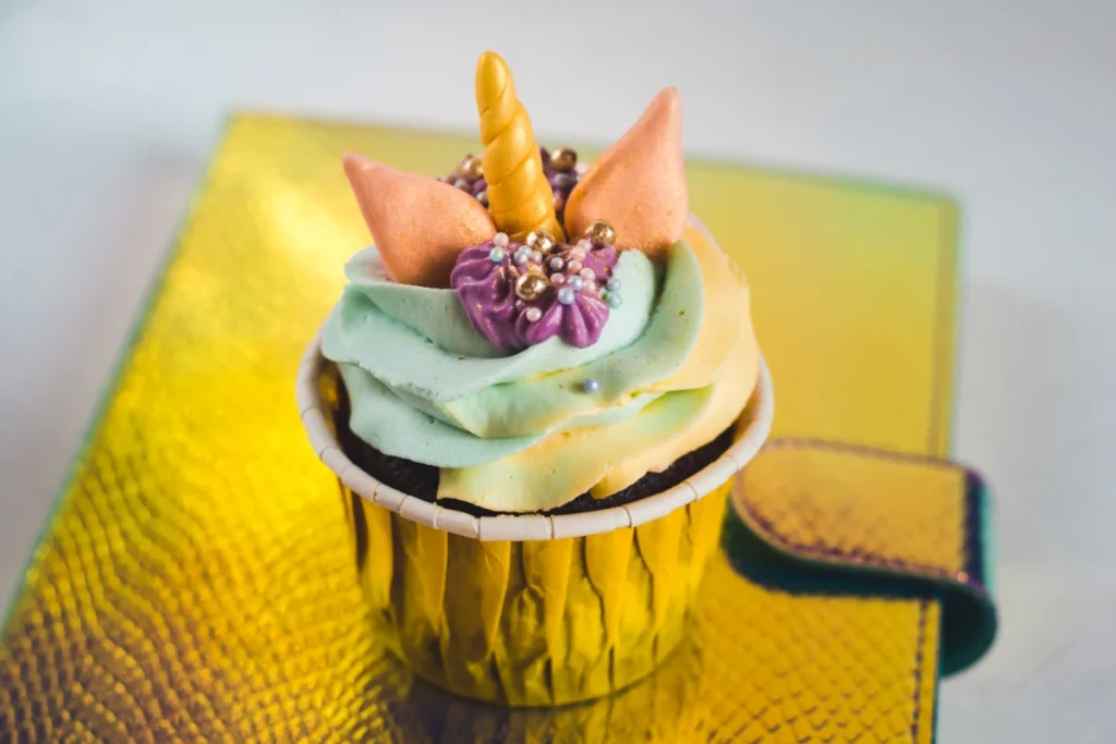 A single unicorn cupcake with a shiny gold wrapper sits on a yellow reflective surface. It is decorated with a golden horn, pastel green and yellow frosting, and embellished with edible pearls and glitter.