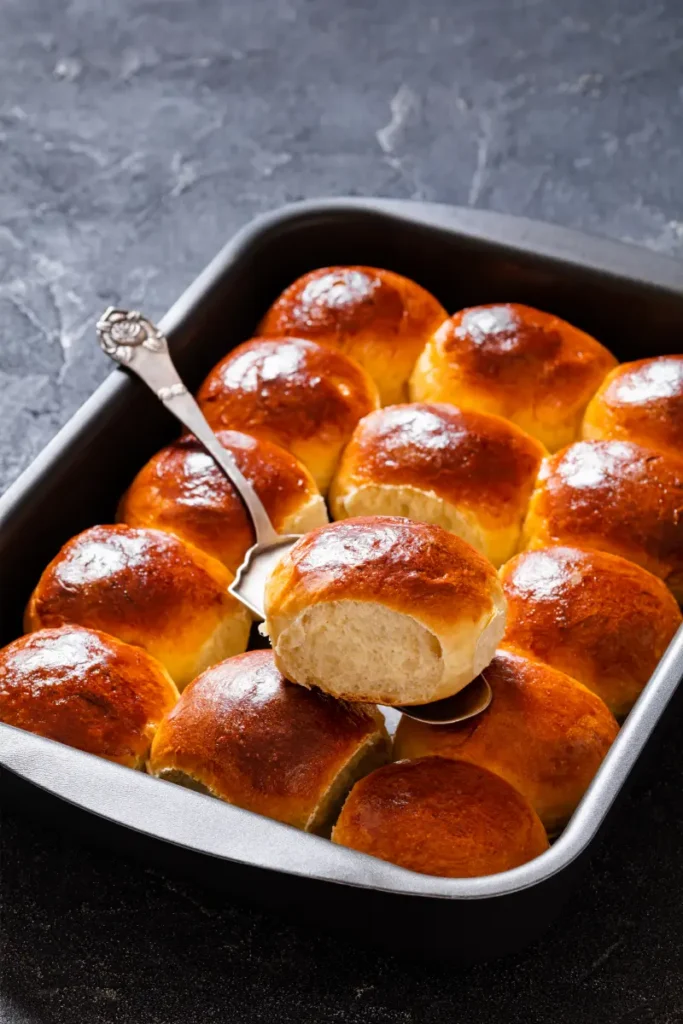 Gluten-free Hawaiian rolls with a shiny, golden crust in a metal baking pan, one roll partially lifted out with a vintage spoon, on a dark surface.