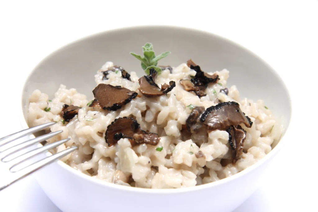 A bowl of truffle risotto garnished with fresh herbs and topped with slices of black truffle, presented on a white background.