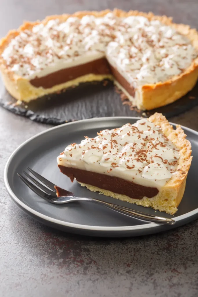 A slice of haupia pie with a creamy white and chocolate layer, topped with whipped cream and chocolate shavings, on a dark grey plate with a fork, alongside the rest of the pie.