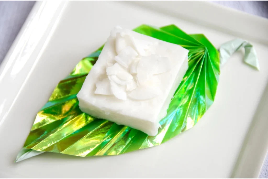 A block of haupia, a traditional Hawaiian coconut milk-based dessert, presented on a green ti leaf with a metallic sheen, placed on a white rectangular plate.