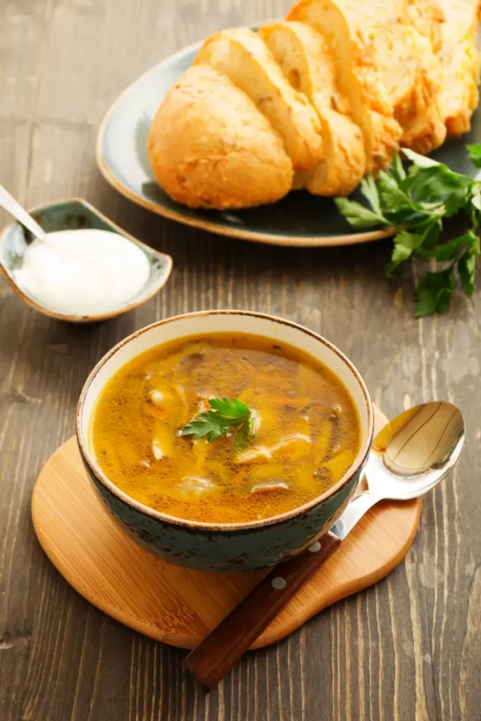 A bowl of oyster mushroom soup garnished with parsley on a wooden board, accompanied by sliced bread on a plate and a small dish of sour cream, with a spoon on the side.