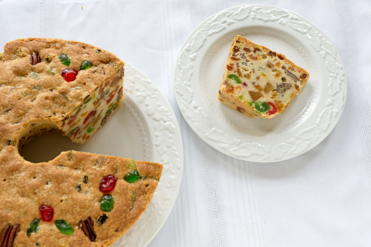 A whole fruitcake with colorful candied fruits and nuts on a white embossed plate, with a single slice placed on a smaller matching plate, all set against a white tablecloth background.