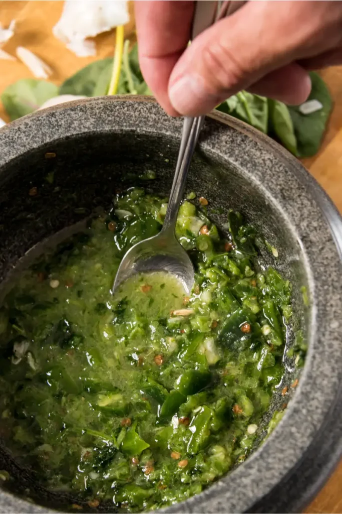 Hand stirring cilantro chimichurri sauce with a spoon in a stone mortar, with garlic cloves and basil leaves in the background.