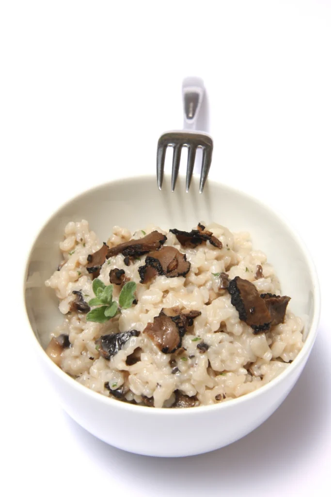 Close-up view of truffle risotto in a white bowl with a fork, topped with black truffle pieces, against a white background.