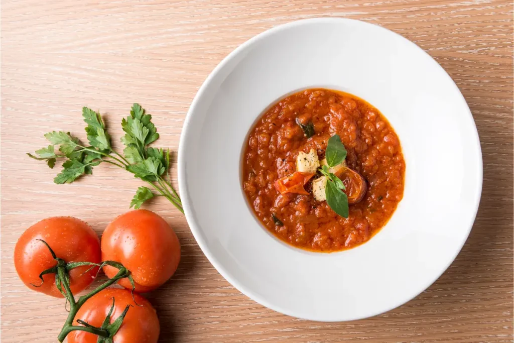 A bowl of robust tomato gravy on a white plate, garnished with a basil leaf and cubes of cheese, accompanied by fresh parsley and vine tomatoes on a wooden surface.