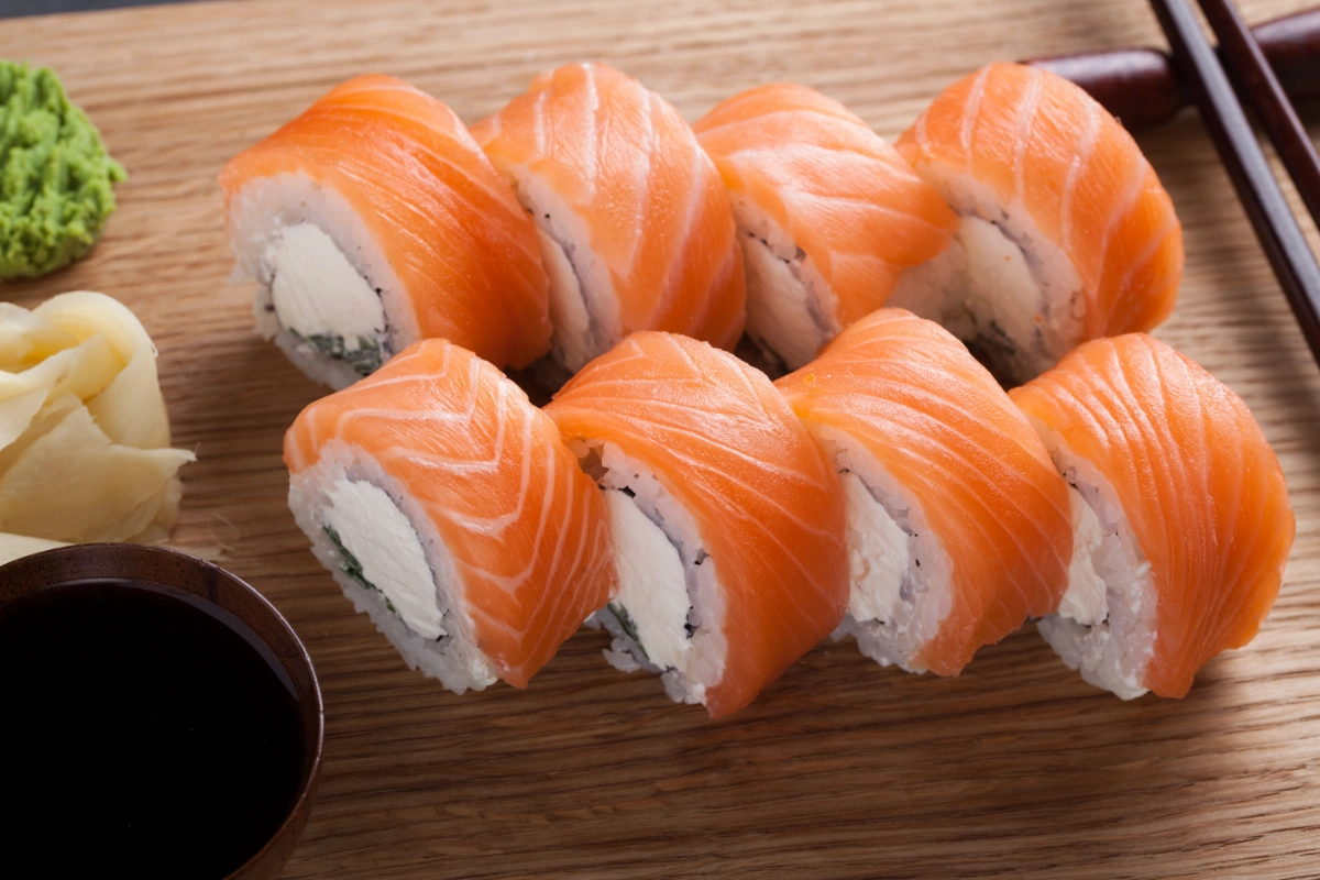 A row of sake sushi, featuring slices of vibrant salmon draped over rolls of rice and cream cheese, served with wasabi, pickled ginger, and soy sauce on a wooden surface.