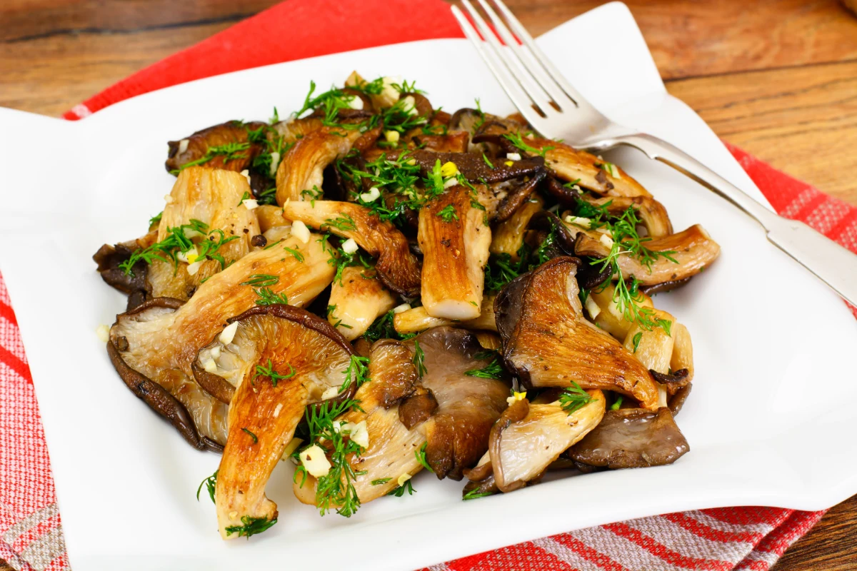 Sautéed oyster mushrooms garnished with fresh herbs on a white plate, with a fork beside it, placed on a table with a red and white checkered napkin.