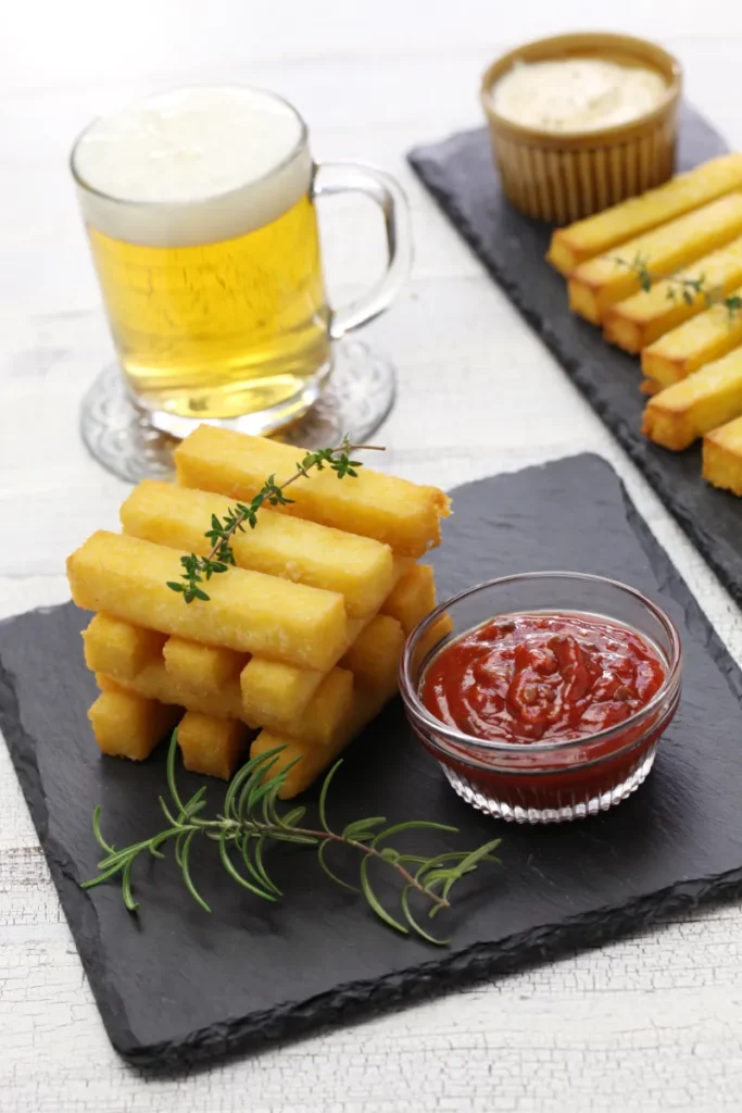 Stacked fried polenta sticks on a black slate board, accompanied by a small bowl of tomato sauce and fresh thyme, with a mug of beer and a bowl of white sauce in the background.