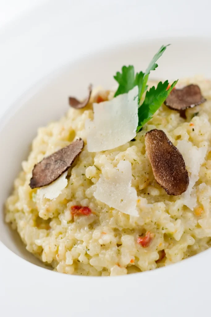 Close-up of a creamy truffle risotto with shaved Parmesan, black truffle slices, and a fresh parsley leaf garnish in a white bowl.