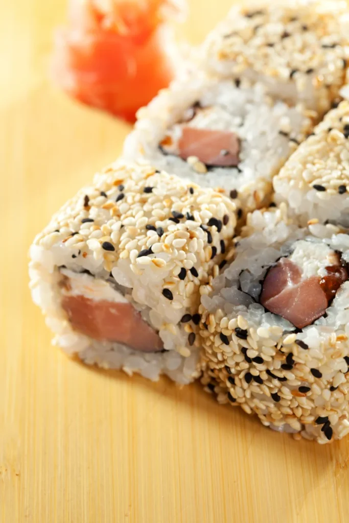 Close-up of Alaska sushi rolls coated with black and white sesame seeds, filled with rice, raw salmon, and a hint of nori, with pickled ginger in the background, on a bamboo mat.