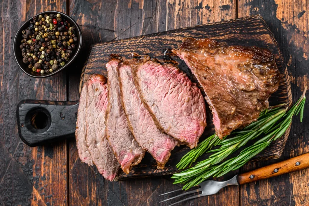 Sliced sirloin tip steak on a wooden cutting board with a sprig of rosemary and a bowl of peppercorns.