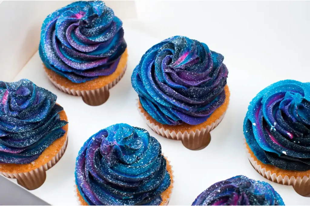 A collection of cupcakes with vibrant galaxy-themed frosting in blue and purple, sprinkled with glitter, presented on a white surface.