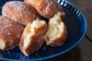 A plate of sugar-coated malasadas, with one split open to show the fluffy interior, on a blue ribbed plate.