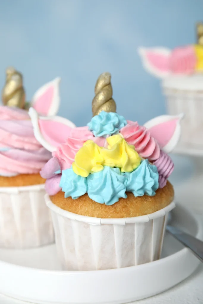 Close-up of a unicorn cupcake with golden horn and ears, frosted with vibrant blue, pink, and yellow cream, showcased on a white cake stand against a blue background.