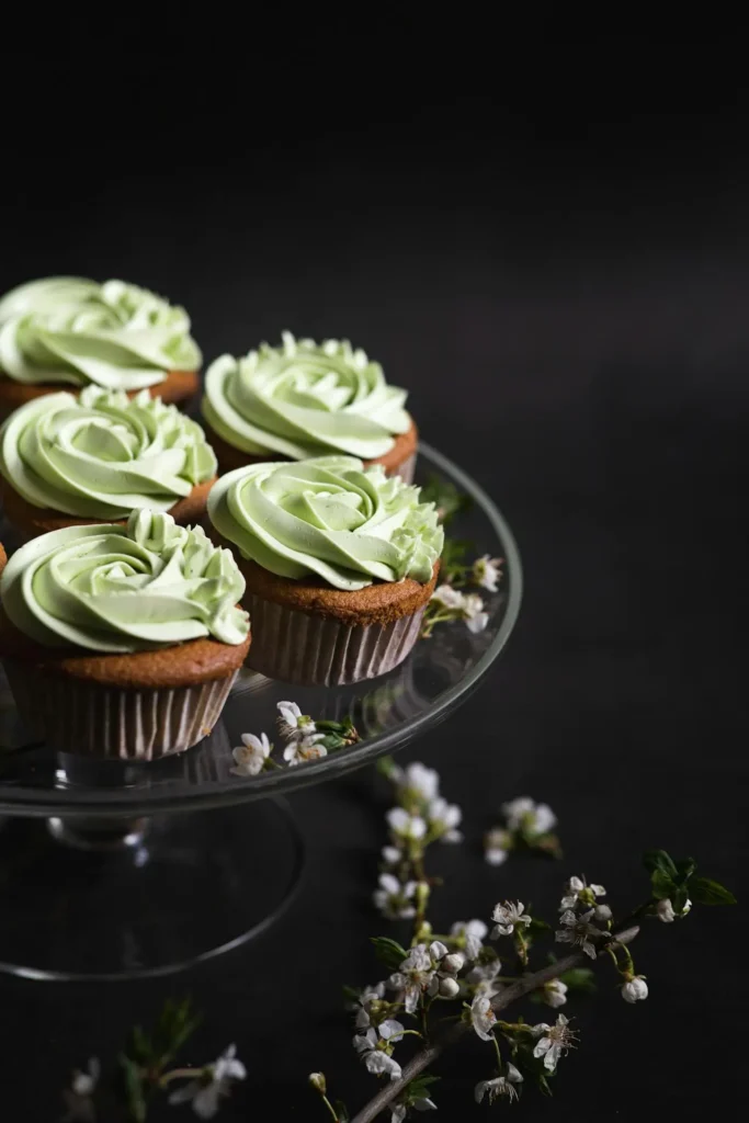 Pistachio cupcakes with swirls of green frosting resembling roses, displayed on a glass cake stand, with white spring blossoms scattered around, set against a dark background.