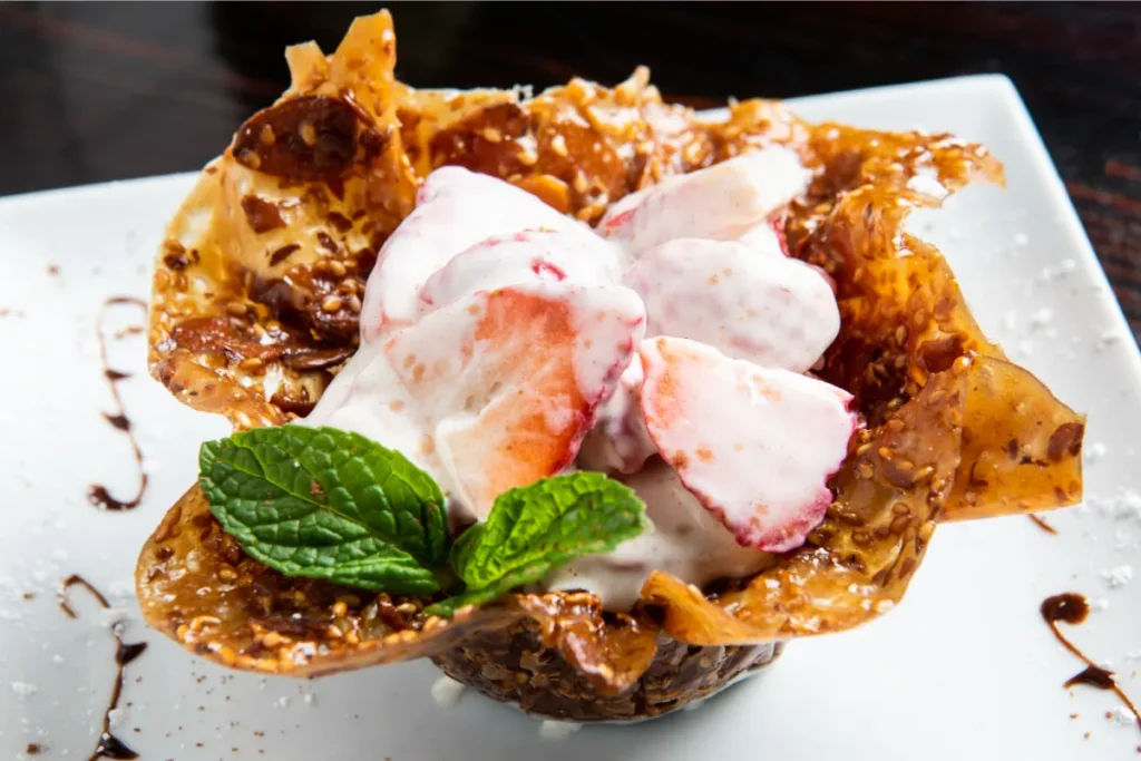 A close-up of a dessert featuring creamy strawberry slices on an almond tuile shell with a mint leaf garnish and artistic chocolate sauce drizzle on a square white plate.