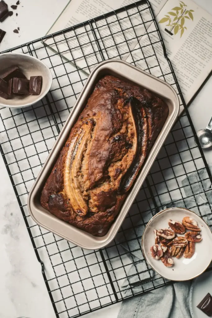 Vegan banana bread in a loaf pan with a caramelized banana on top, on a cooling rack beside ingredients of pecans and dark chocolate on a marble surface.