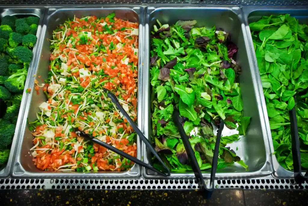 Salad bar selection featuring a mixed vegetable salad with diced tomatoes, alongside trays of mixed leafy greens and fresh spinach, with serving tongs.