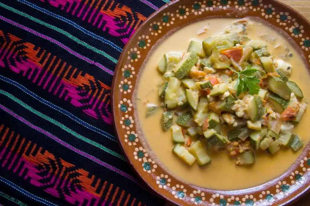 A plate of Calabacitas, a traditional Mexican dish with zucchini, cheese, and tomato, on a colorful woven textile, garnished with fresh cilantro.