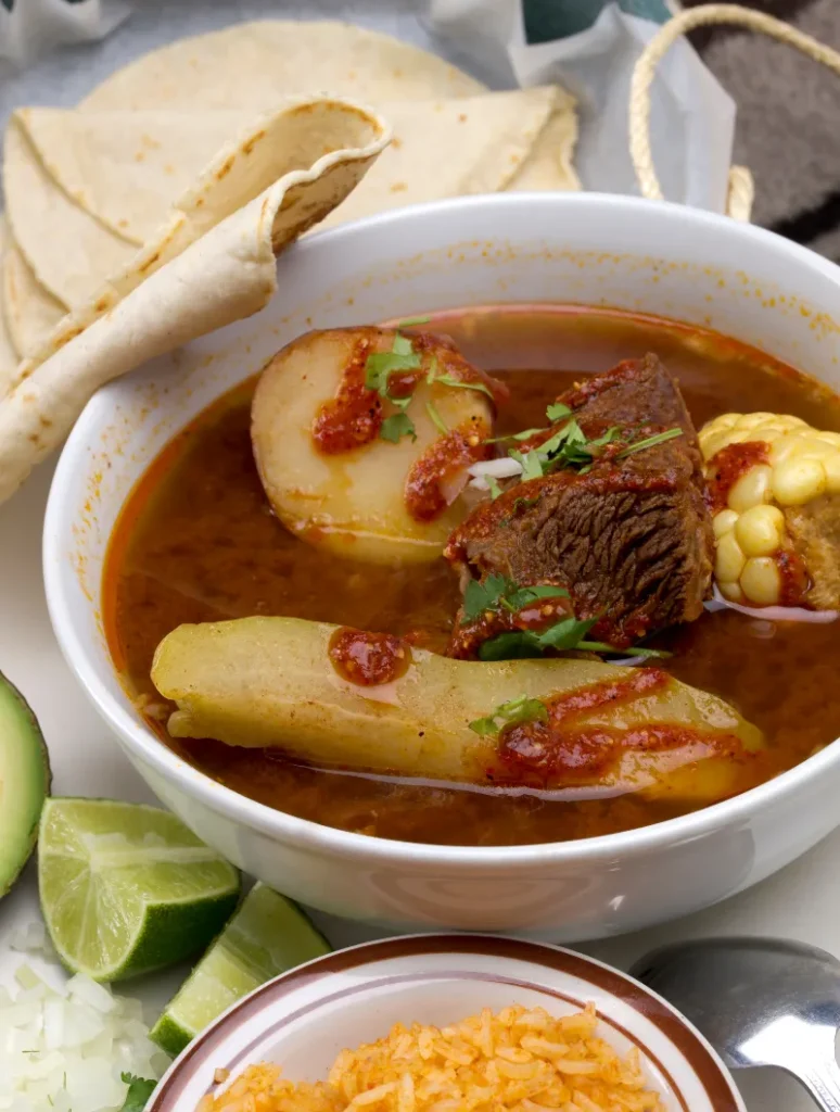 Bowl of Caldo de Res with beef, corn, and potatoes in a red broth, accompanied by sides of diced onions, lime wedges, rice, avocado slices, and folded tortillas.