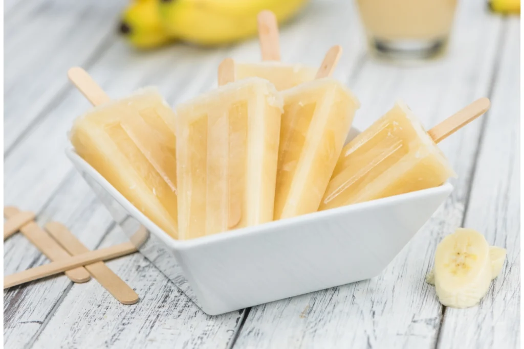 A white bowl filled with homemade banana popsicles on wooden sticks, with a single banana slice in the foreground and whole bananas and a glass of banana smoothie slightly blurred in the background.