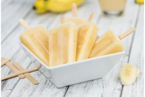 A white bowl filled with homemade banana popsicles on wooden sticks, with a single banana slice in the foreground and whole bananas and a glass of banana smoothie slightly blurred in the background.