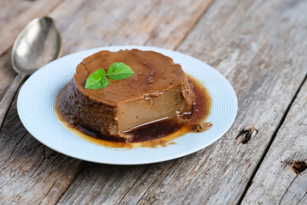 A coffee flan on a blue-striped white plate, garnished with a green basil leaf, with caramel sauce and a spoon on a rustic wooden table.