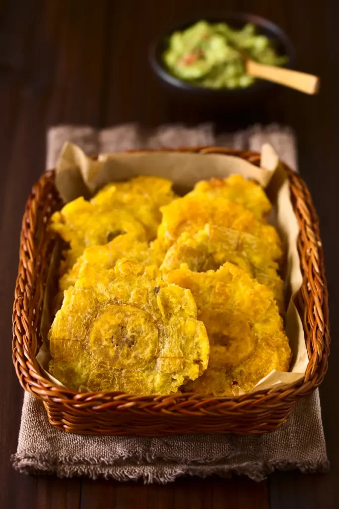 A basket filled with yellow patacones, served on a burlap cloth, with a bowl of guacamole and wooden utensils in the blurred background.