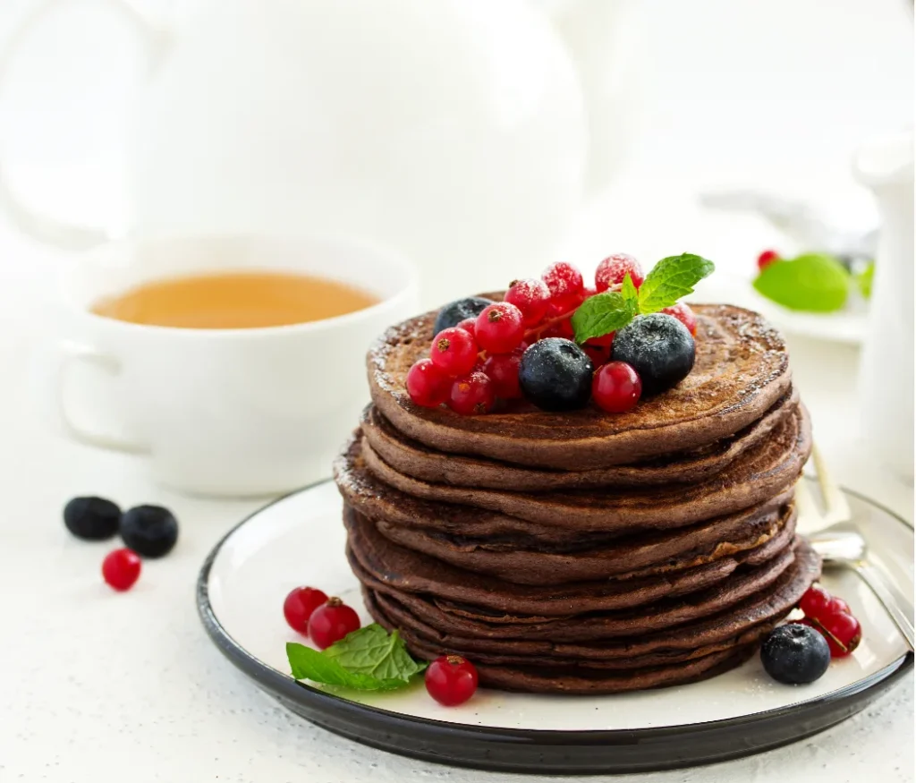 A plate of chocolate pancakes topped with a mix of red currants and blueberries, dusted with powdered sugar, and accented with a fresh mint leaf. A cup of tea is in the background.