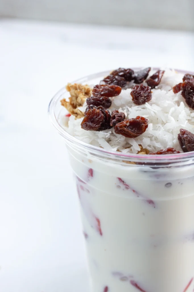 A parfait with layers of strawberries and cream, topped with granola and raisins, presented in a clear glass against a white background.