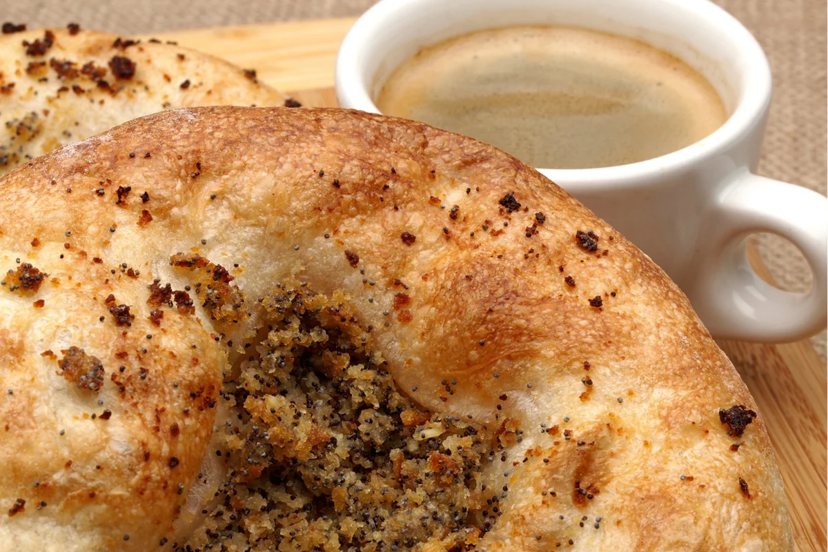 Close-up of a bialy bagel sprinkled with poppy seeds, accompanied by a cup of coffee in the background.