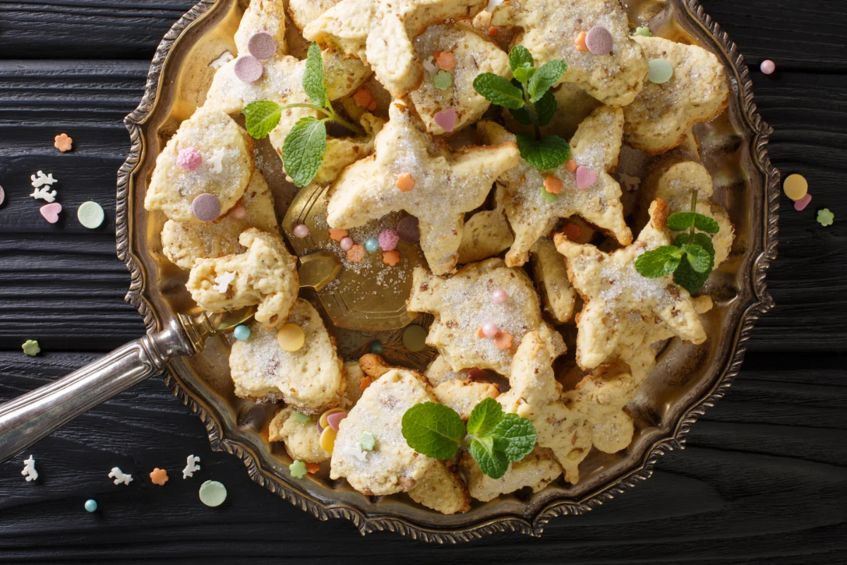 A plate of biscochitos, traditional star and moon-shaped cookies sprinkled with sugar, adorned with pastel confetti and fresh mint leaves, on an ornate silver tray against a dark wood background.