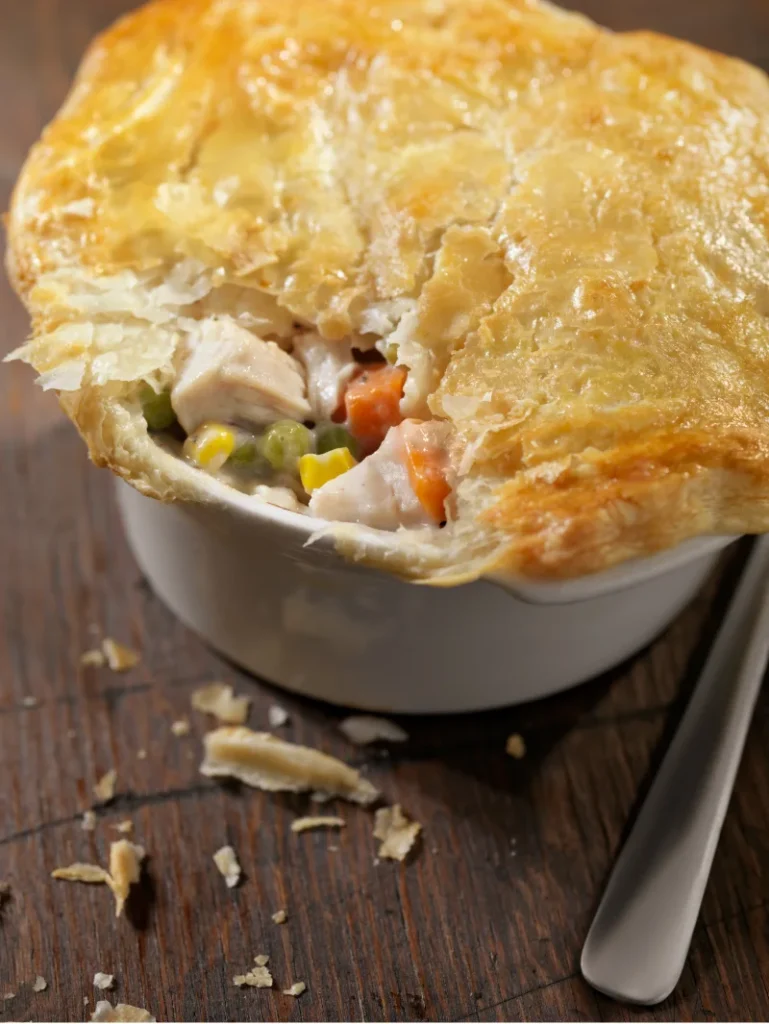 Close-up of a Bisquick Chicken Pot Pie with its pastry top partially lifted, exposing the creamy chicken and vegetable filling inside, accompanied by a spoon, on a wooden surface with crumbs scattered around.