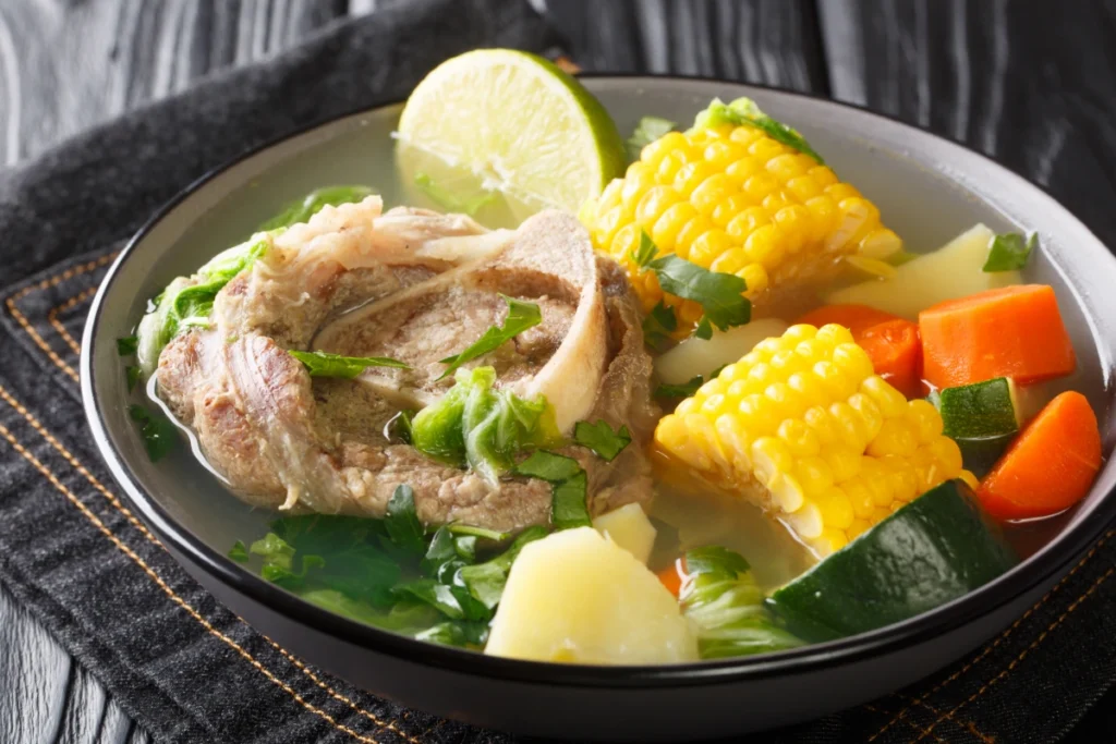 A bowl of Caldo de Res, a traditional Mexican beef soup, featuring a large piece of beef with bone, chunks of corn on the cob, diced carrots, potatoes, and zucchini, garnished with lime and cilantro.